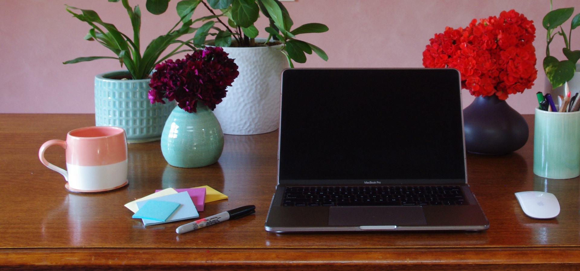 A desk with a laptop, vases of flowers, stationary and a mug