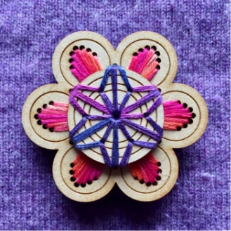 A wwoden brooch featuring embroidery in purples, pinks and oranges